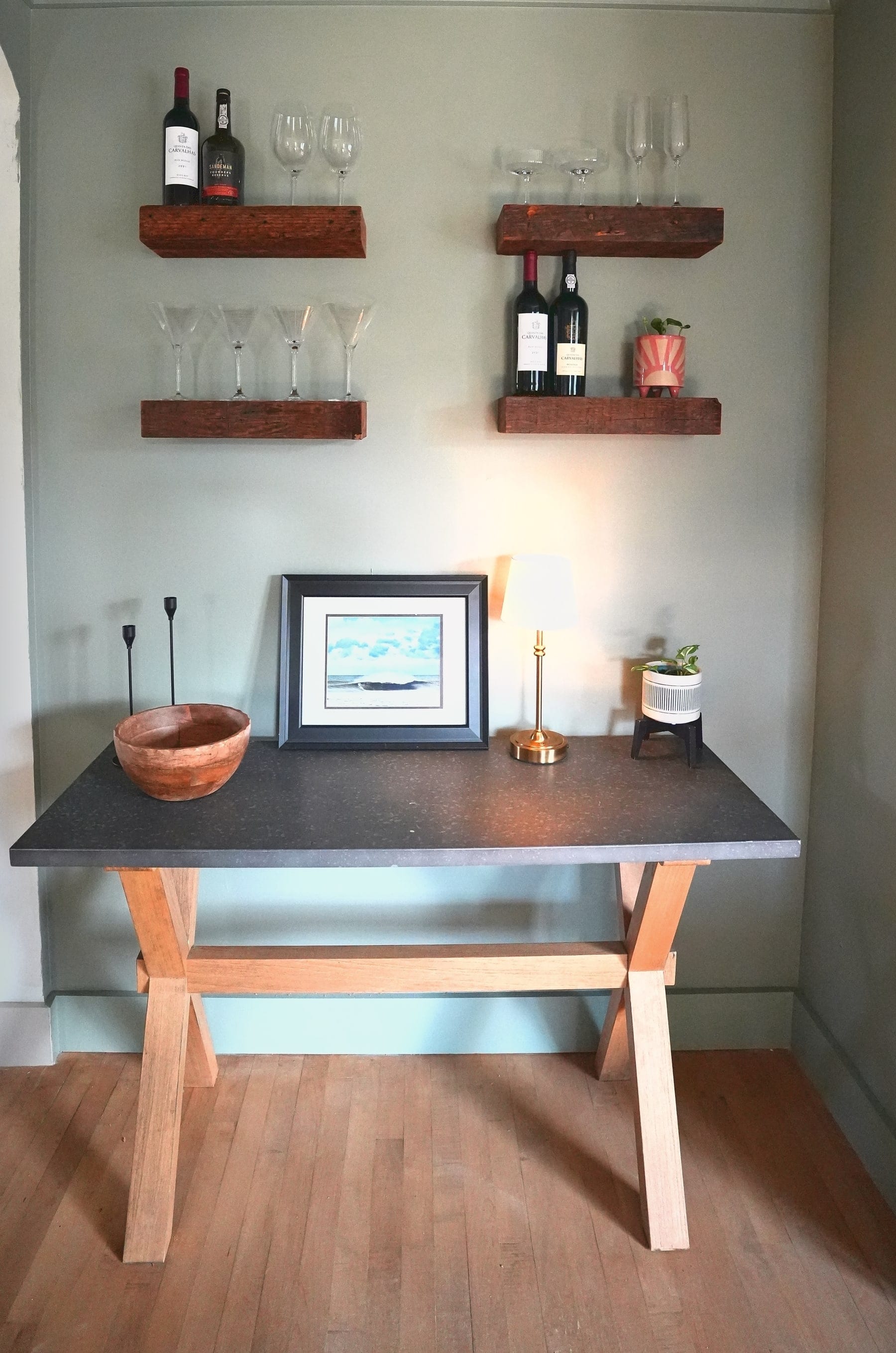 Four wood floating shelves holding wine bottles and glasses above a bar console with a dark top and X-frame wood legs