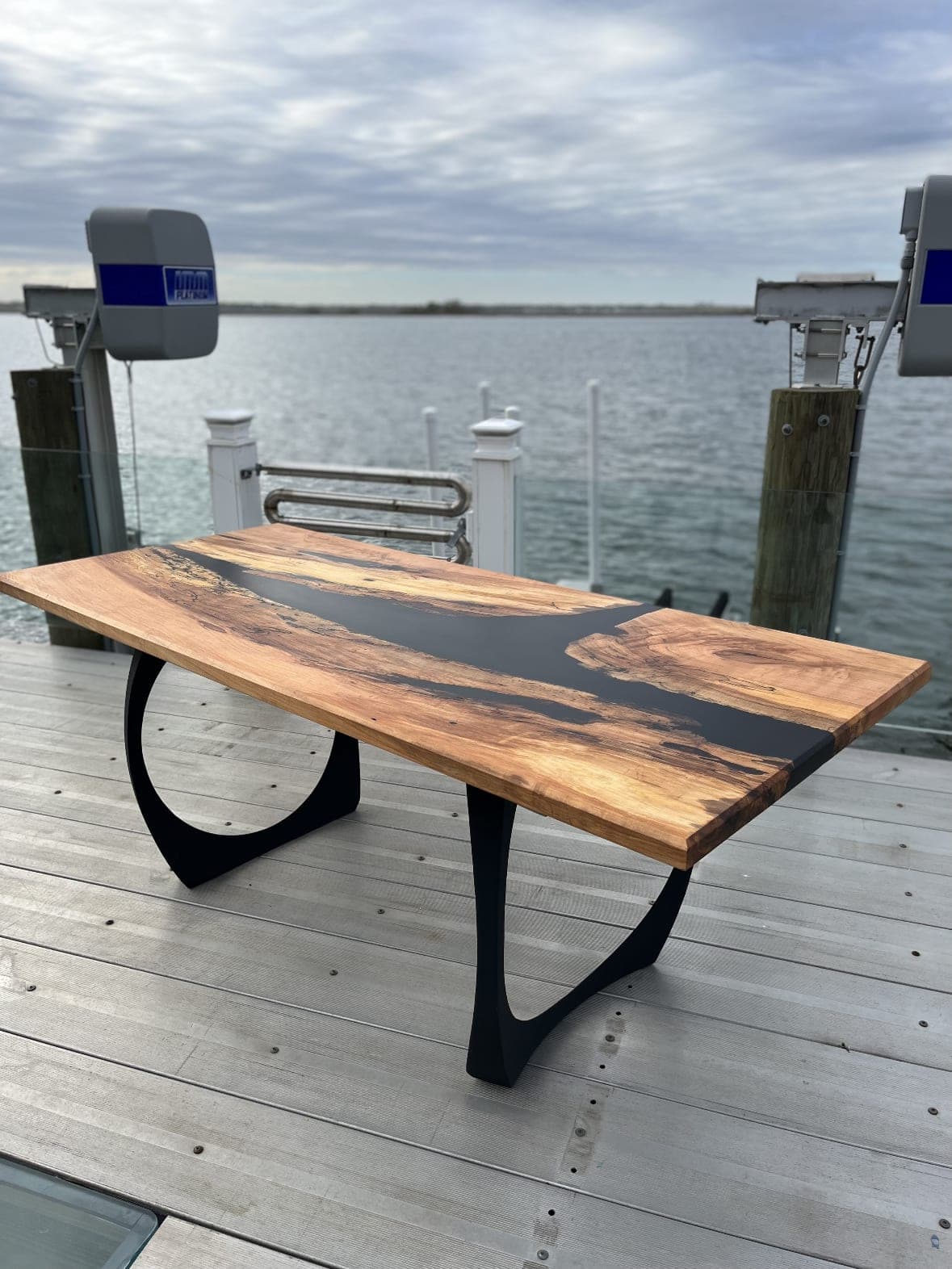 Live-edge table with a dark resin river on black steel legs, on a dock by the water
