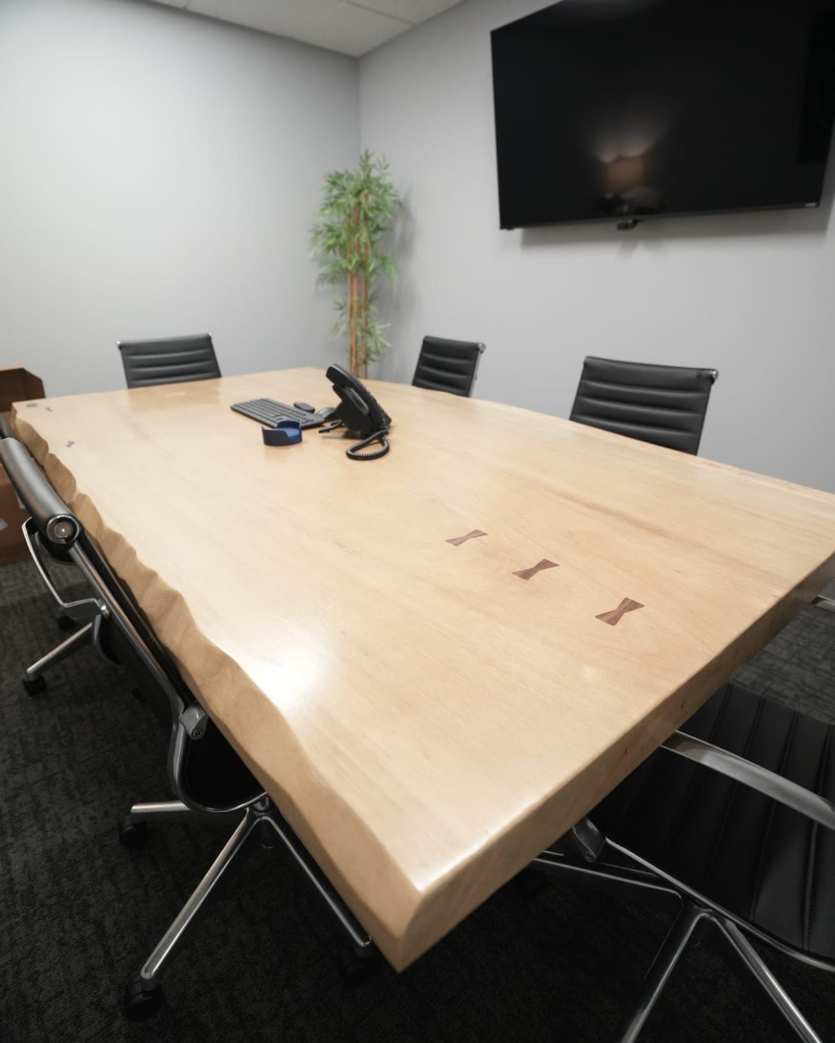 Light live-edge conference table with a row of dark bowtie inlays, surrounded by black office chairs